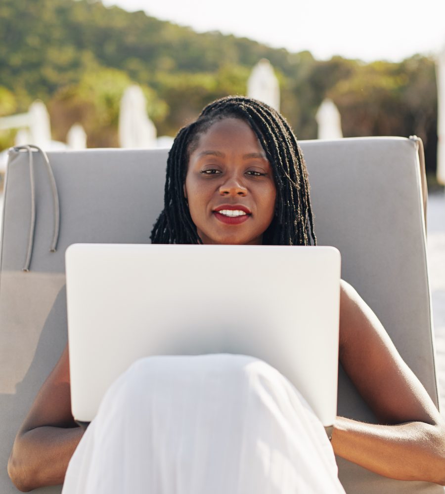 Young Black female digital nomad sitting on chaise-longe on sandy beach and working on laptop