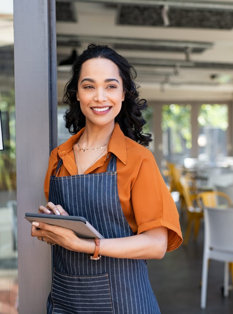 Portrait of happy waitress standing at restaurant entrance and holding digital tablet. Happy young latin woman owner in apron standing at coffee shop entrance, leaning while waiting clients. Successful cafe business owner looking away with copy space.
