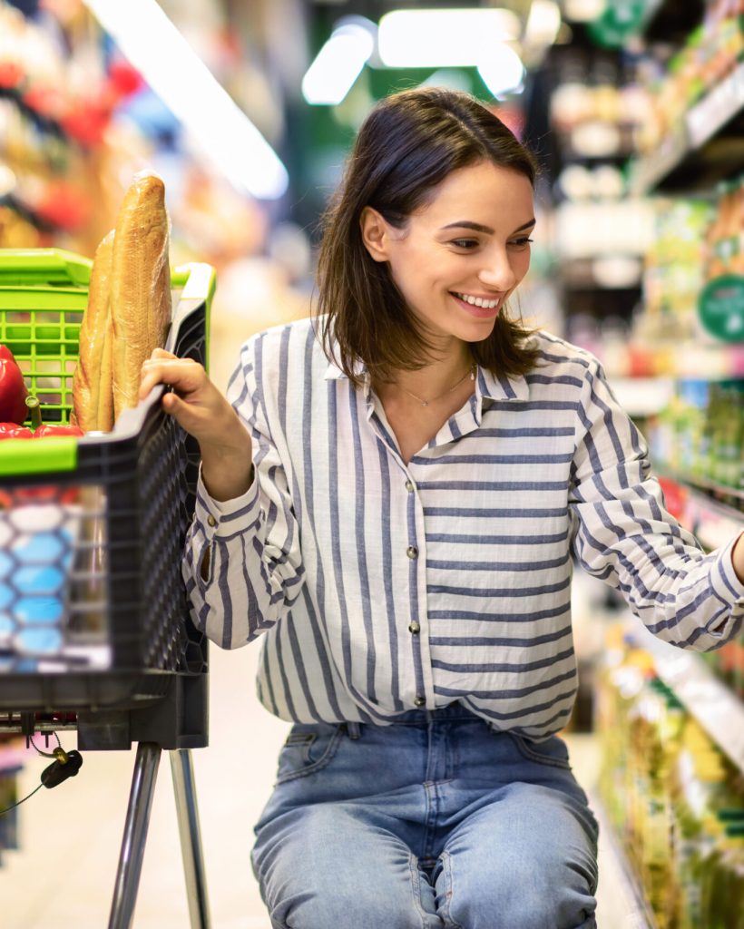 At The Supermarket. Portrait Of Smiling Millennial Lady With Shopping Cart In Wholesale Market Buying Groceries Food Nutrition, Taking Products From Shelves In Store, Low Angle View
